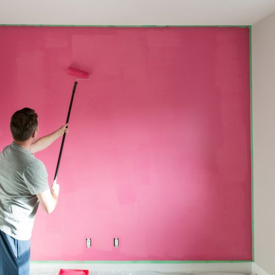 A man painting a wall in pink color with a paint roller.