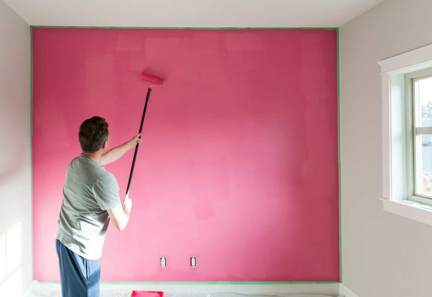 A man painting a wall in pink color with a paint roller.