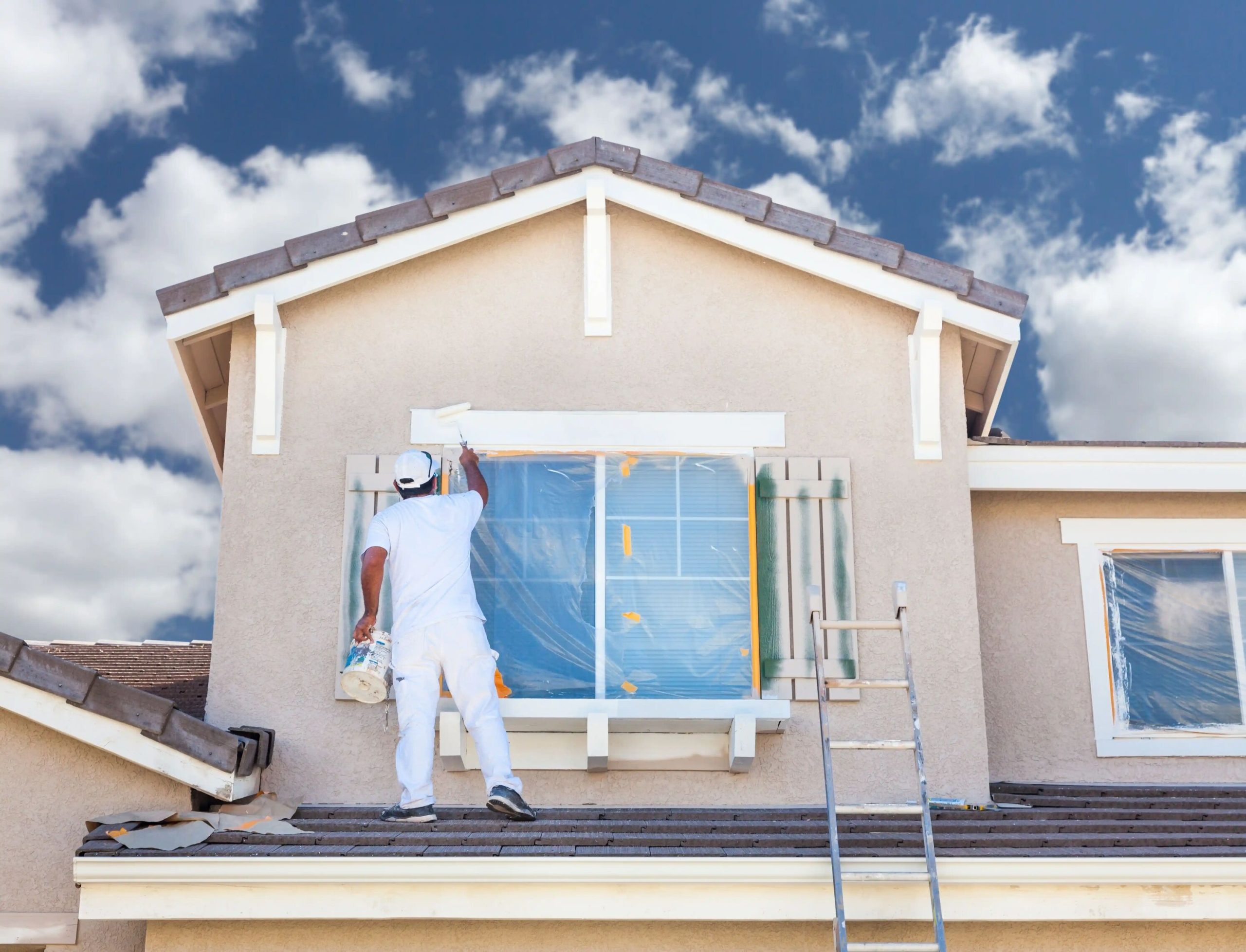 Painter standing on a roof applying white exterior wall paint around a window for long-lasting protection, with the window covered for precision.