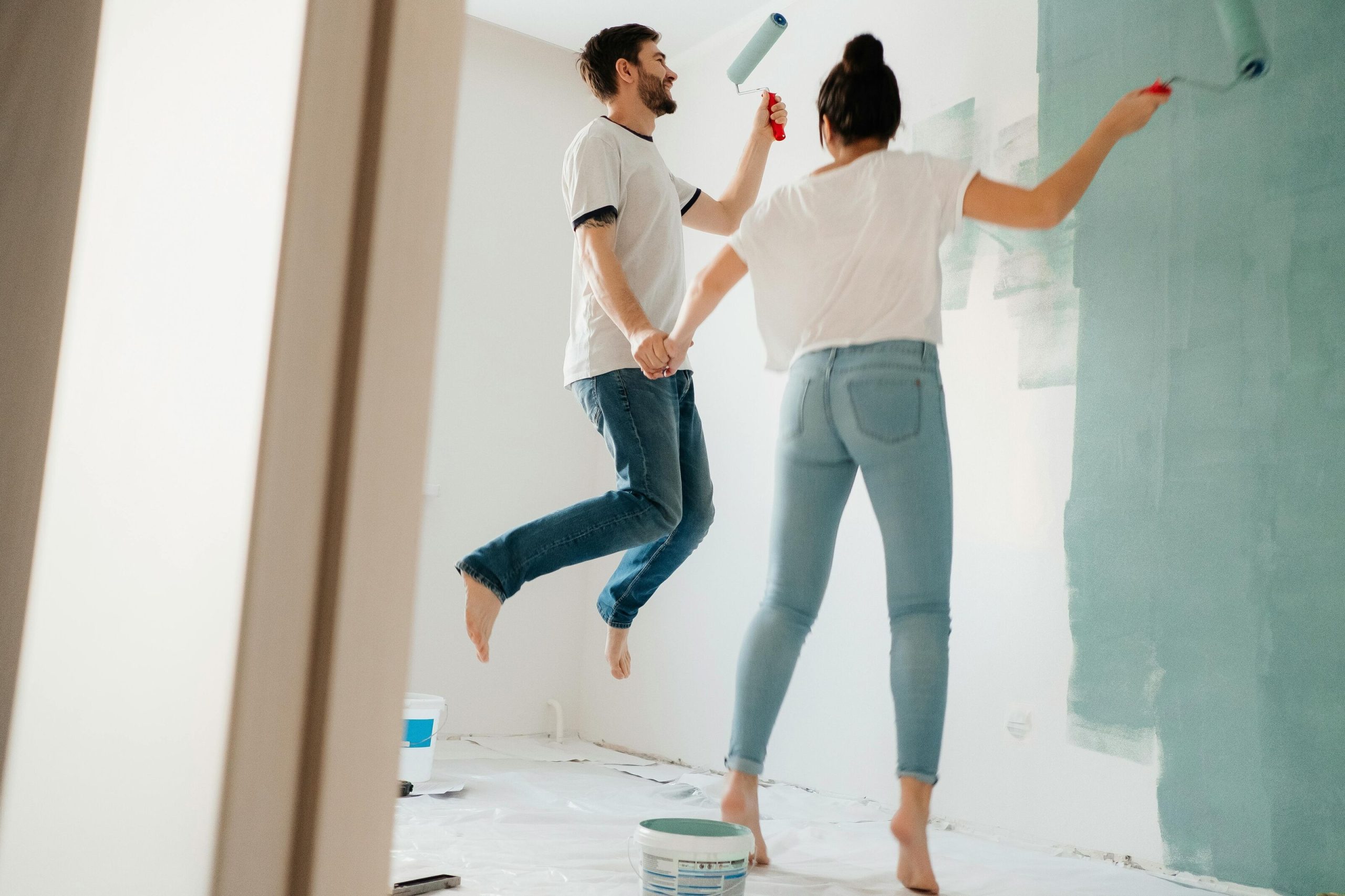 Couple joyfully painting a wall light green with rollers while holding hands, symbolizing sustainable home improvement and eco-friendly living.