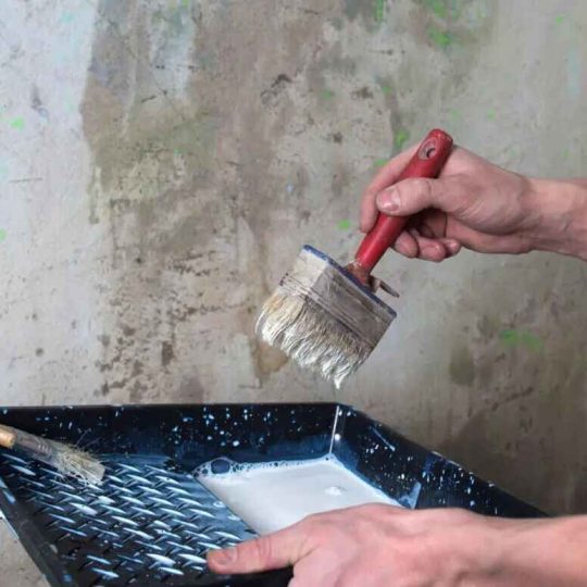 Hand dipping paintbrush into white paint tray during wall preparation.