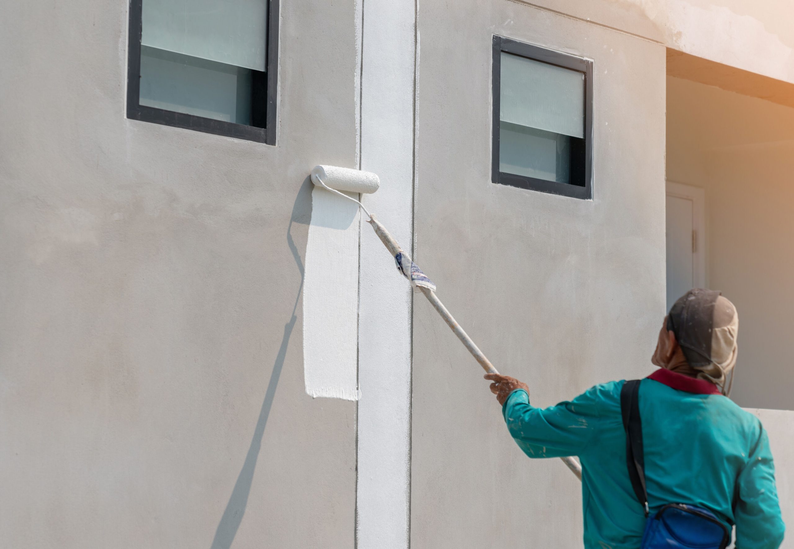 Exterior house painting with roller applying white paint to wall, protecting home surfaces from rain and humidity in a tropical climate.