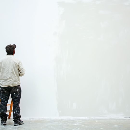 Painter standing on a ladder applying white paint to a large wall, illustrating how to choose the right white shade for Singapore homes without a flat finish.